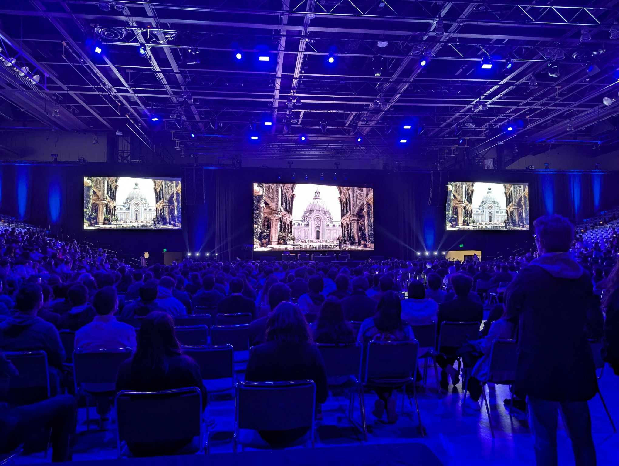 A live audience facing a stage and three large display surfaces in a blue-lit venue.